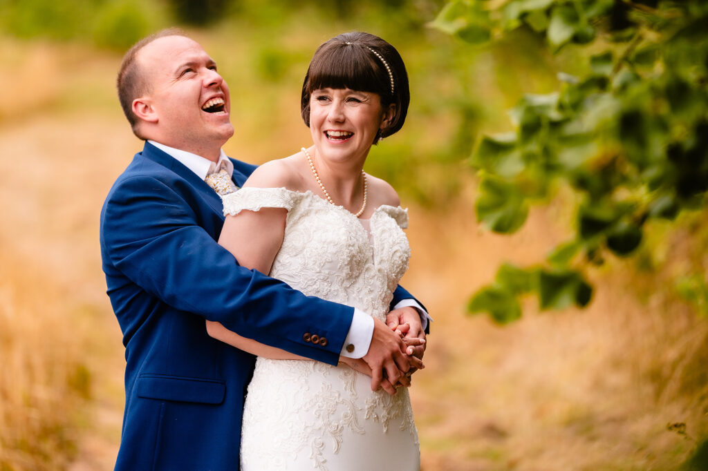 Groom hugging the bride from behind as they both laugh, framed by leaves, Cheshire wedding photographer