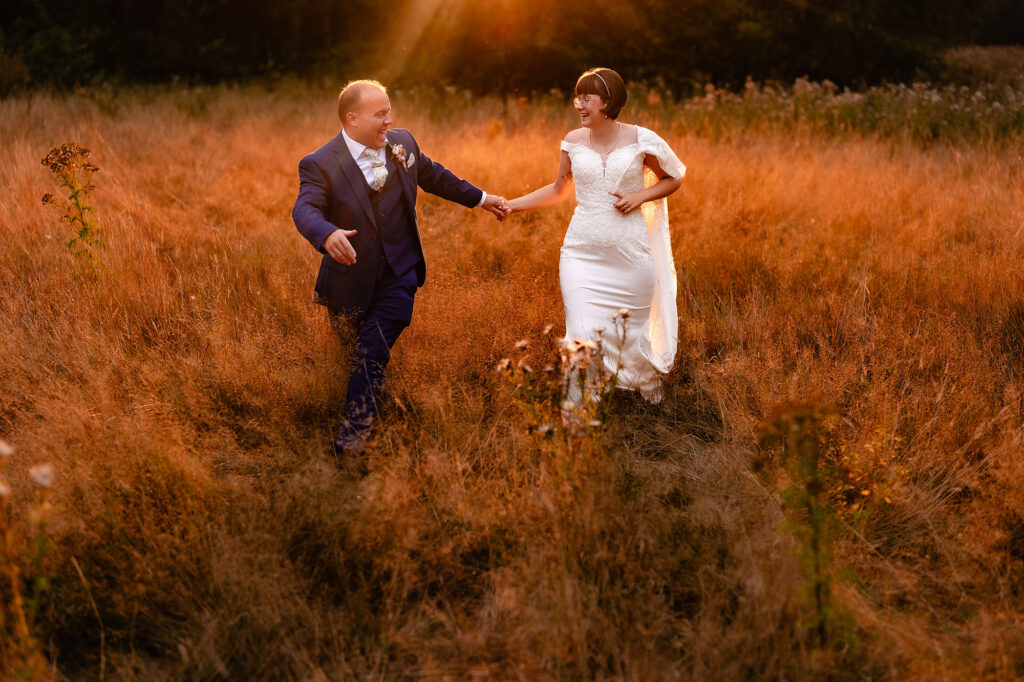 Bride and groom running hand in hand through long grass glowing orange at golden hour at The Oak Tree of Peover